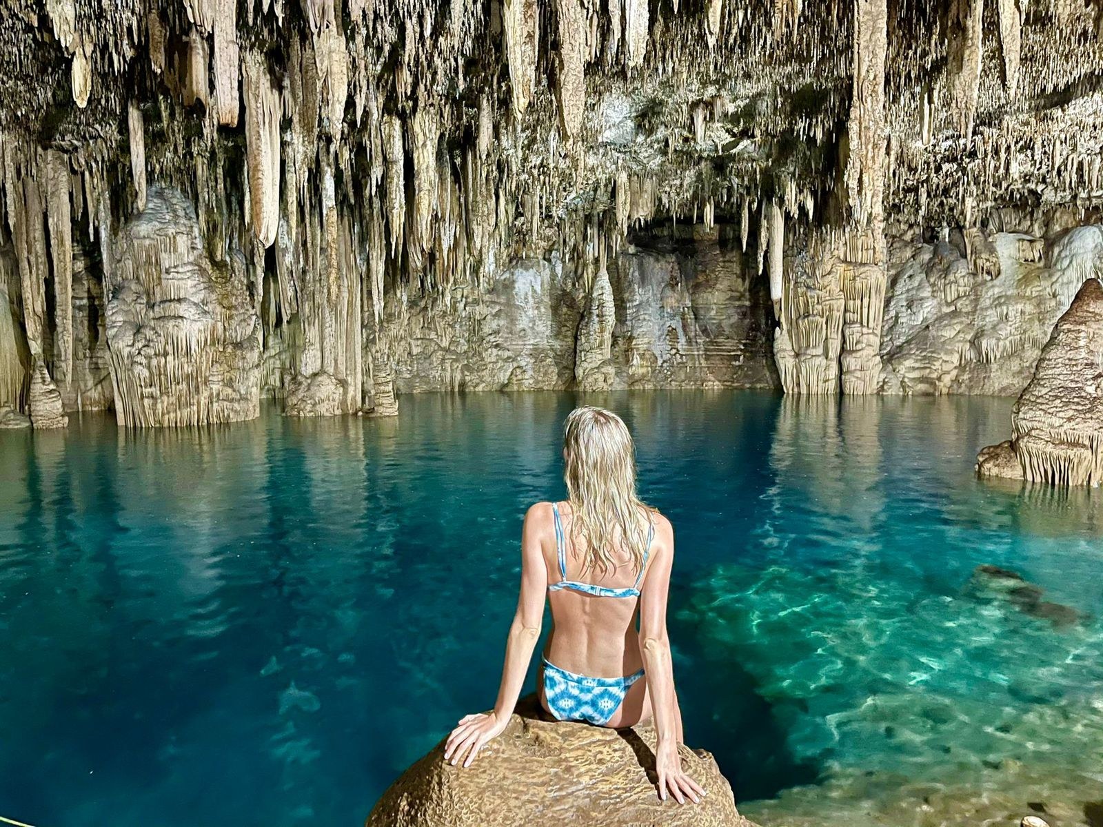 Viajera sentada en una roca frente al agua azul de un cenote de caverna con estalactitas