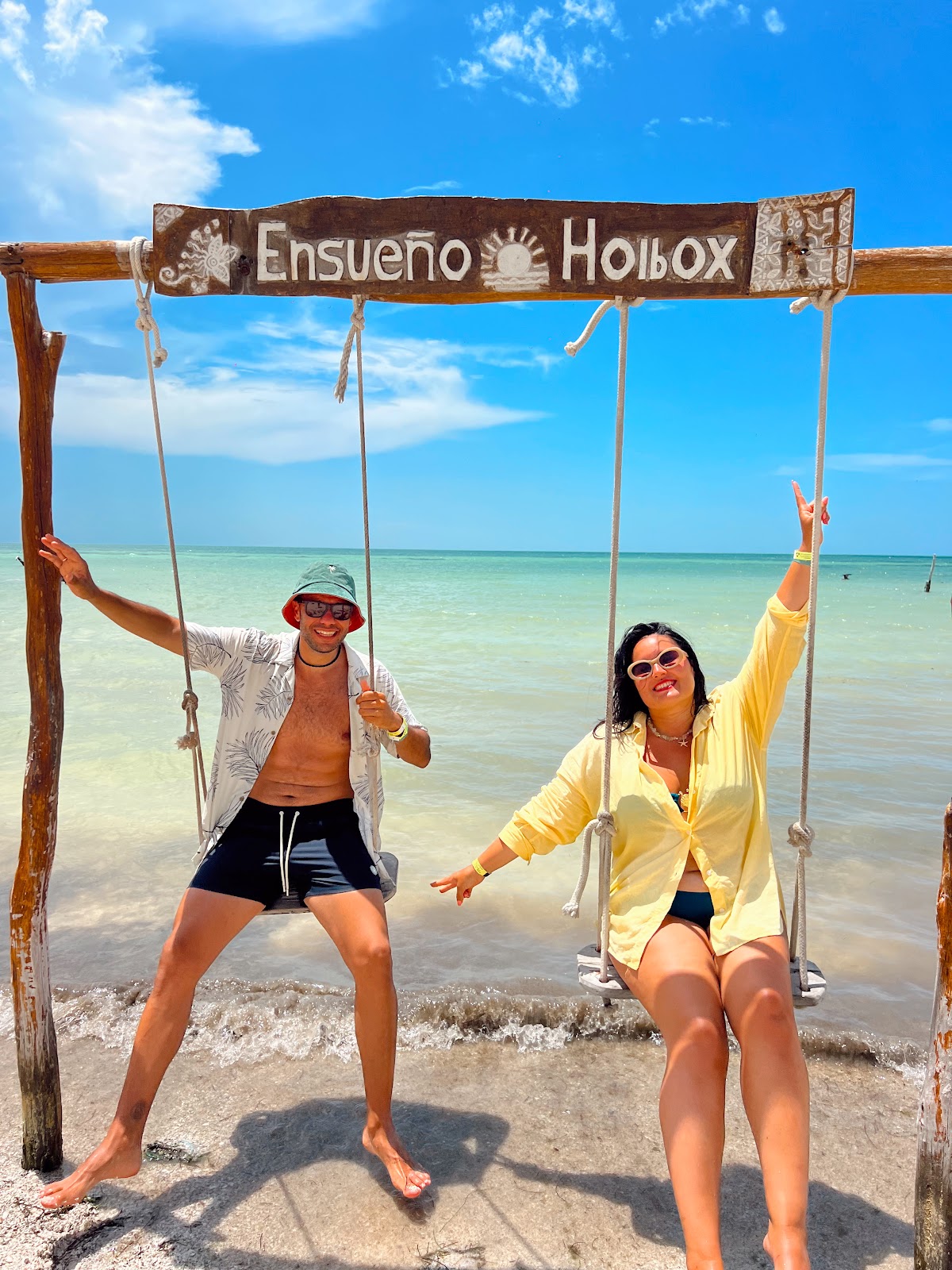Pareja de viajeros en el columpio de madera «Ensueño Holbox», con el mar de Isla Holbox detrás