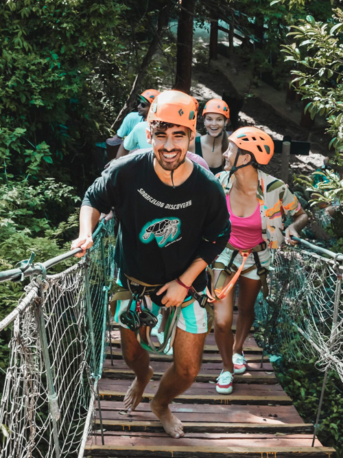 Grupo de viajeros cruzando un puente colgante en la selva con cascos y arneses